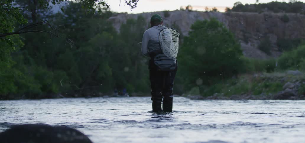 A man with a hat a net and a fishing rod fly fishing in a river