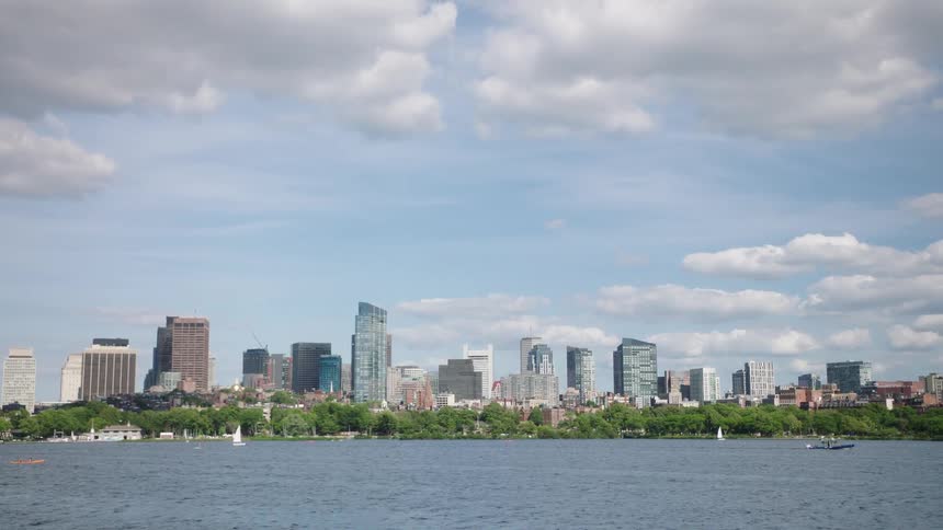 A large city with many skyscrapers sits in the distance from a large body of water with boats floating about on a cloudy day