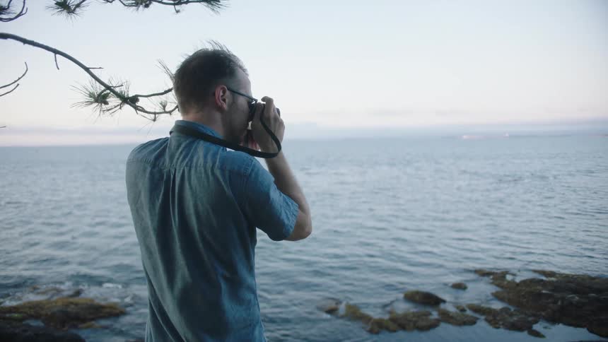 A man wearing eyeglasses stands at an elevated point overlooking an ocean and takes pictures with a camera hung around his neck