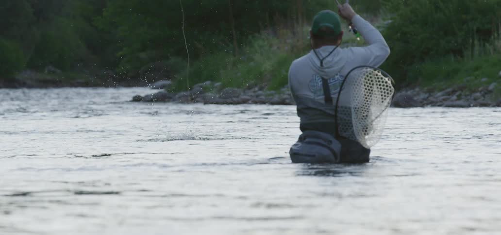 A fisherman standing waist deep in a river pulls back his fishing pole causing a fish to thrash around above the water