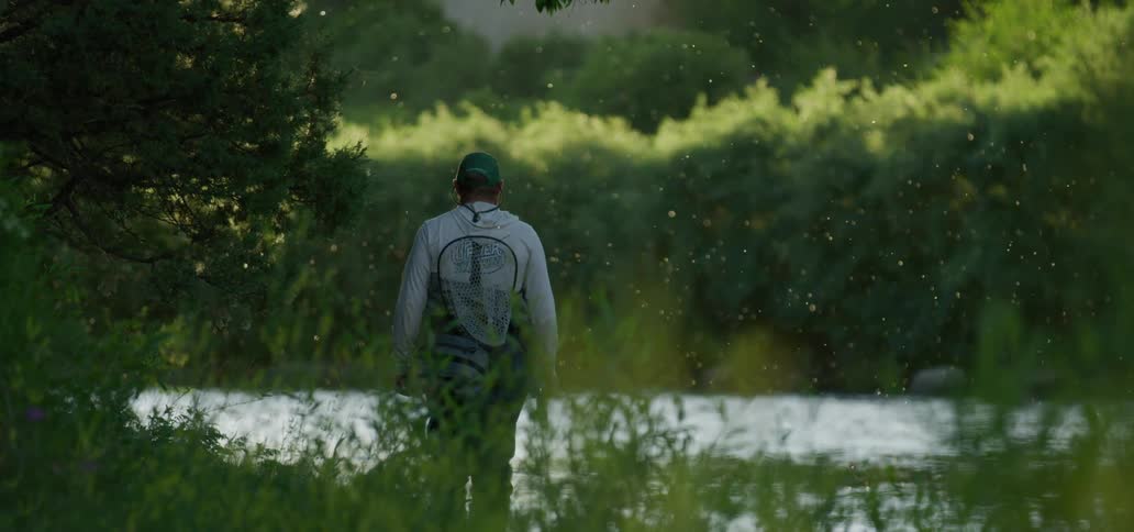 A man with a hat a net and a fishing rod walking by a river