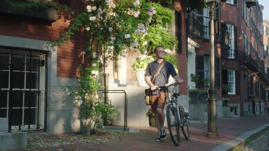 A man pushes a bicycle along a red brick sidewalk in a city neighborhood during the day