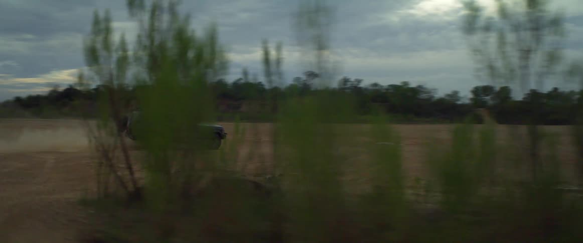 A vehicle driving and raising dust with trees the sky and clouds in the background