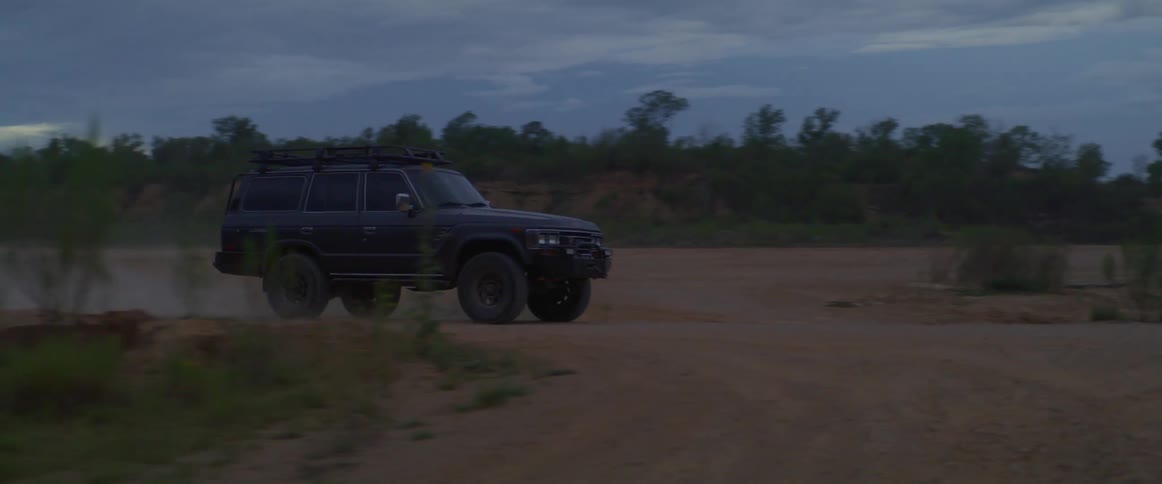 A vehicle travels along a dirt road through the countryside on a cloudy day