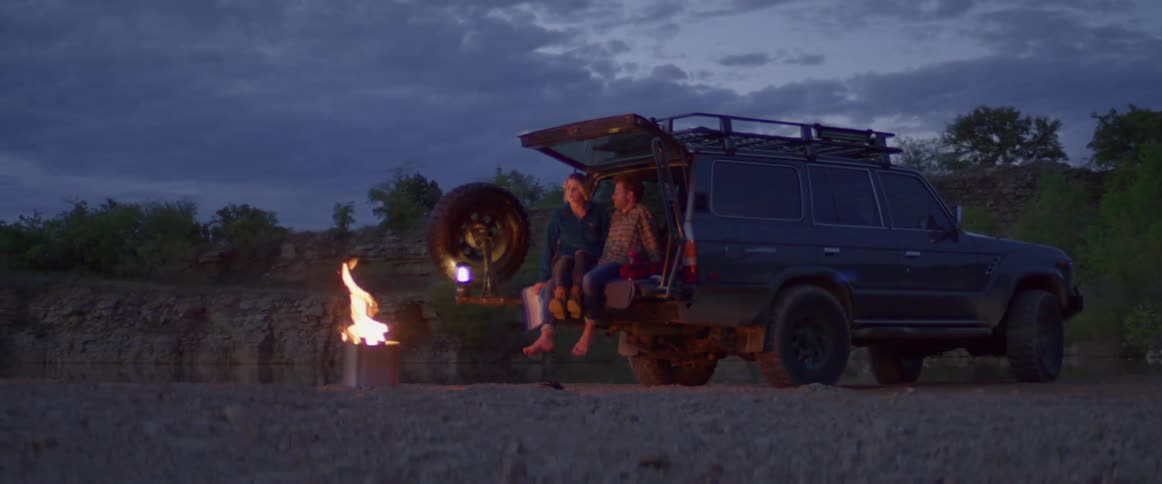 A man and woman sit in the back of their vehicle with a campfire in front of them