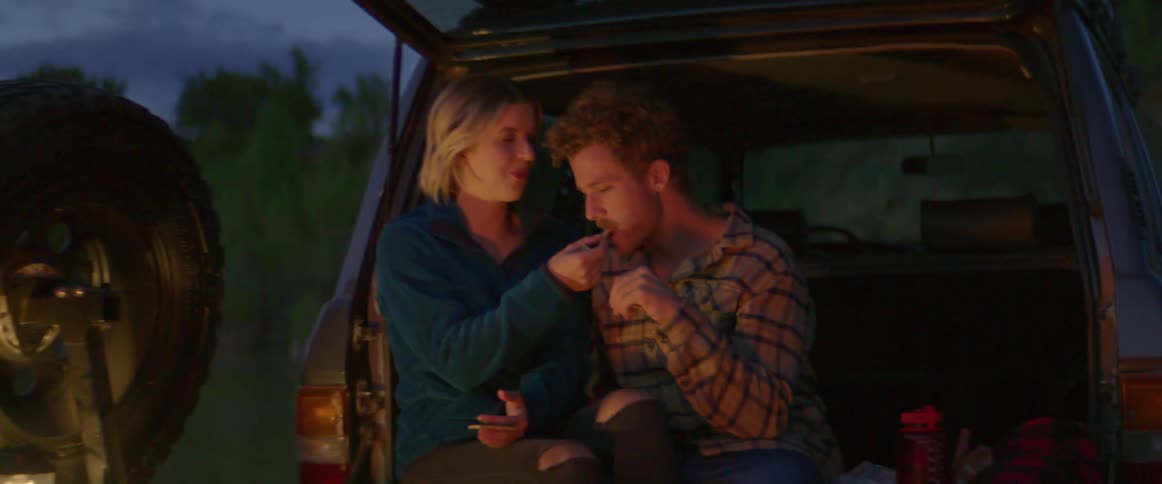 A young couple sit in the back of their vehicle and snack on food near a campfire