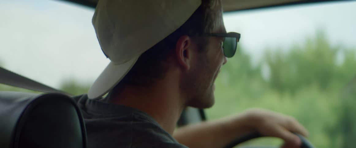 A man driving a car talks happily with his passenger as they travel through the countryside on a cloudy day