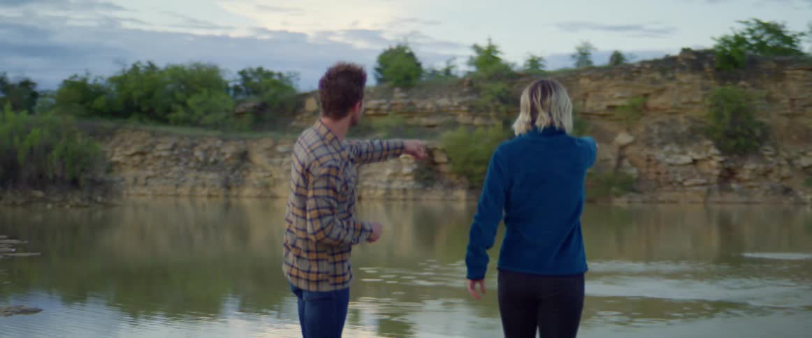 A man and a woman skipping rocks in a creek with trees the sky and clouds in the background