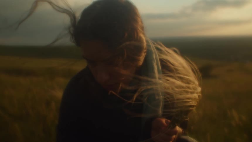 A female crouches down and picks tall grass from a field on a windy day