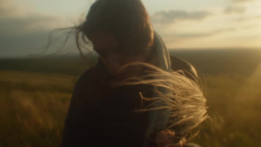 A woman holding silky grass blades with dark black hair is standing on a grassy plain with yellow dried grasses blowing in the wind