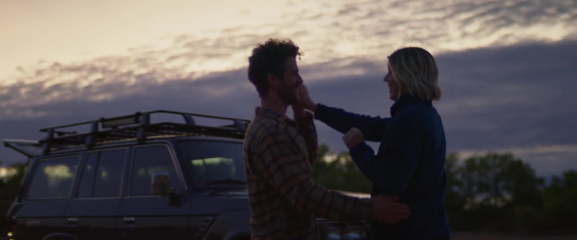A man and a woman dancing with a vehicle trees the sky and clouds in the background