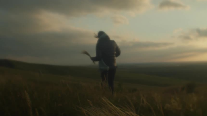 A woman with a coat and scarf walking in a grassy field with the sky and clouds in the background