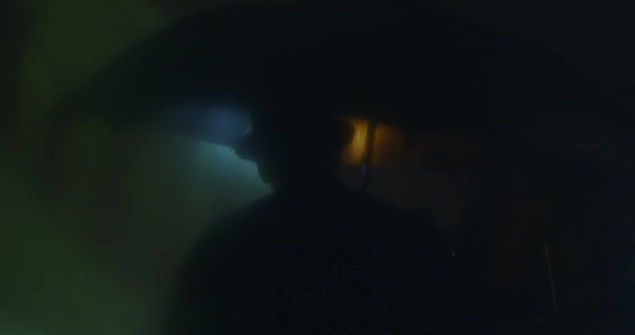 Mans silhouette stands holding umbrella in foggy and shadowy area near dim blue and amber lights