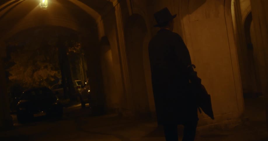 A man wearing a long coat and hat swings an umbrella back and forth as he walks down the hallway of a dim building