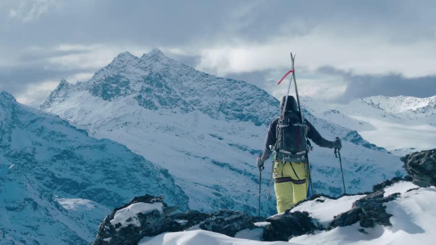 A mountaineer looks out over a mountain valley while standing atop a snowy mountain ridge on a cloudy day