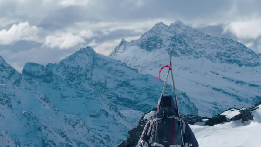 A person with a backpack and skis walking on a snow covered cliff with a mountain range the sky and clouds in the background