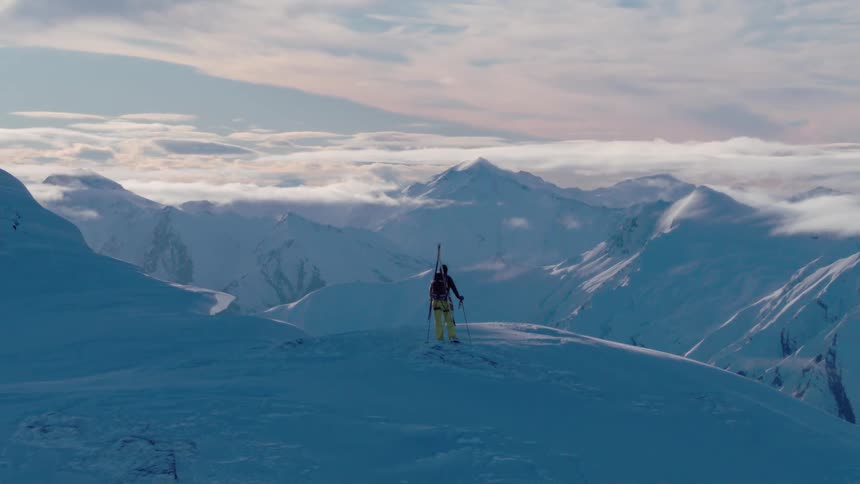 A person with skis standing on a cliff with a mountain range the sky and clouds in the background