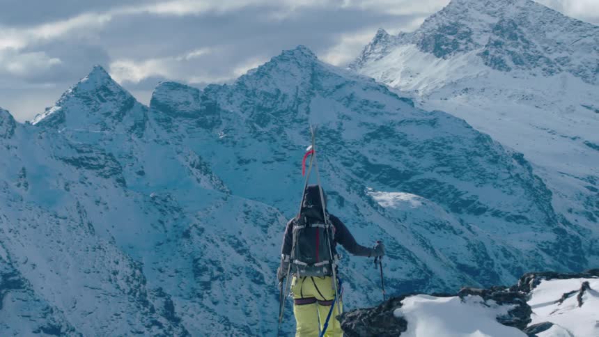 A person wearing yellow and black protective snow gear is standing on a snowy mountain peak looking out at the mountainous icy landscape under a cloudy sky