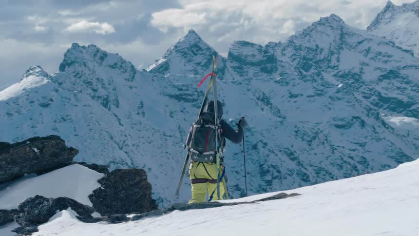 A person with skis on their back hiking up a mountain with snow with a lake in the background