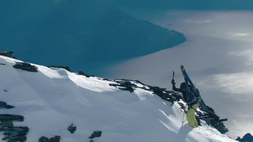A person with skis on their back hiking up a mountain with snow with a lake in the background