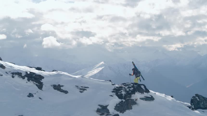 Woman with skis on back hikes up snowy ridge on tall mountain