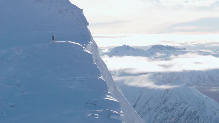 A mountaineer stands high on a snow covered mountain under a cloudy sky
