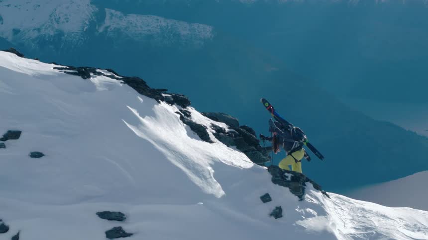A person with skis on their back hiking up a mountain with snow with a lake in the background