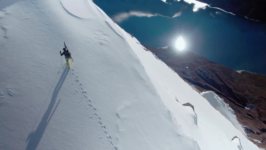 A mountaineer hikes across a snowy mountain slope high above a lake reflecting the sky