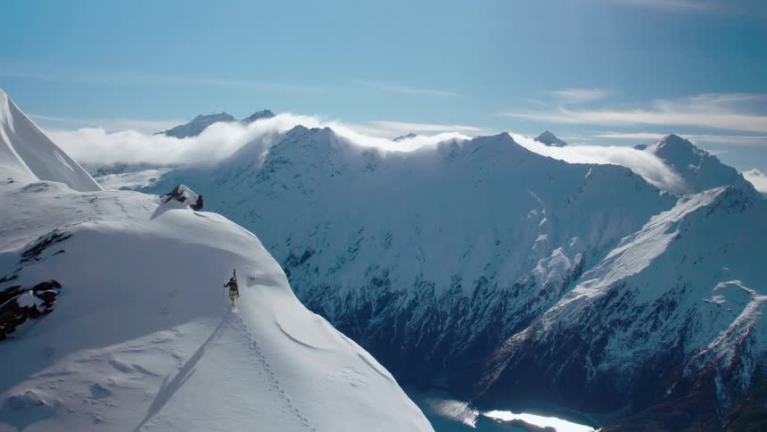 A person with skis on their back walking up a snow covered mountain with the sky and clouds in the background