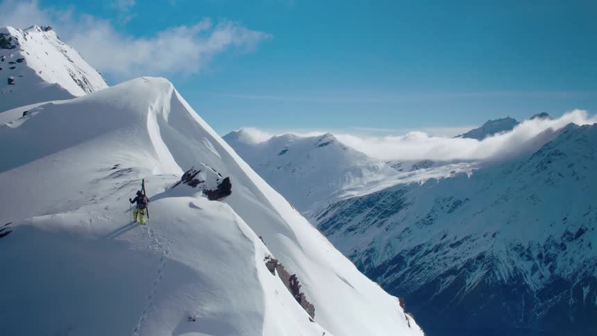 A mountaineer hikes up across a snow covered mountain ridge on a sunny day