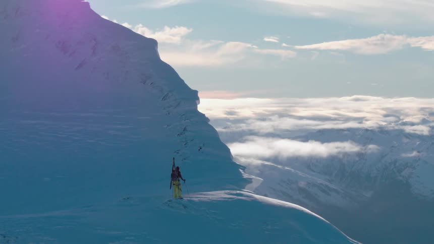A person with skis on their back standing on a mountain covered in snow with the sky and clouds in the background
