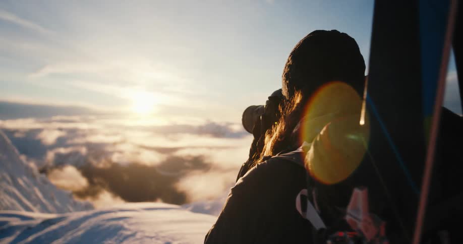 Woman with skis on her back stands on a snowy mountain and takes a photo