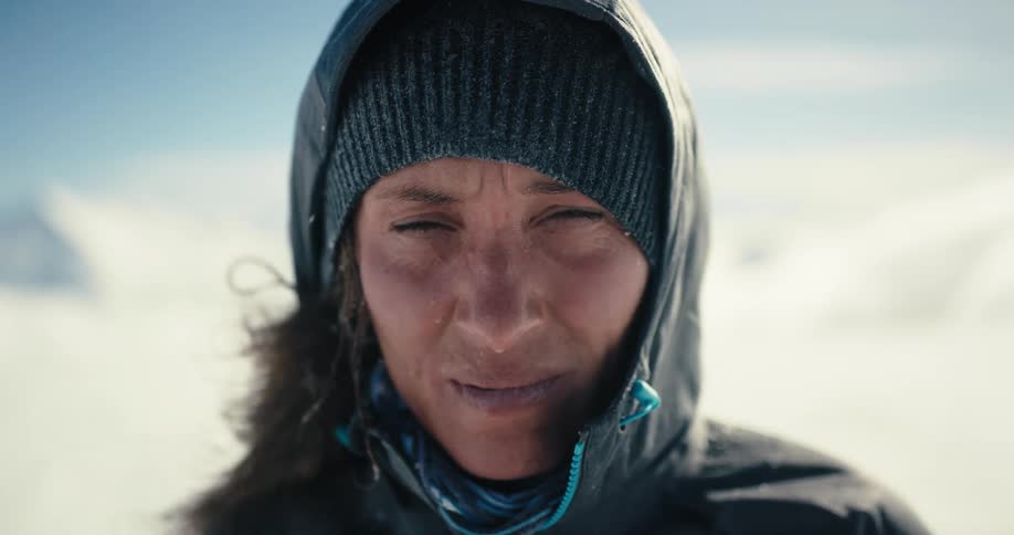 A woman standing outside in the blowing wind turns to look at a snow covered mountain behind her