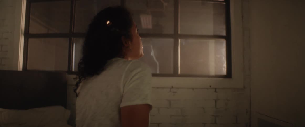 A woman standing in a dark bare room looks out a window
