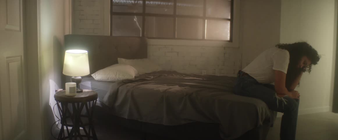 A depressed young woman sits on the edge of a bed while gazing down at the floor