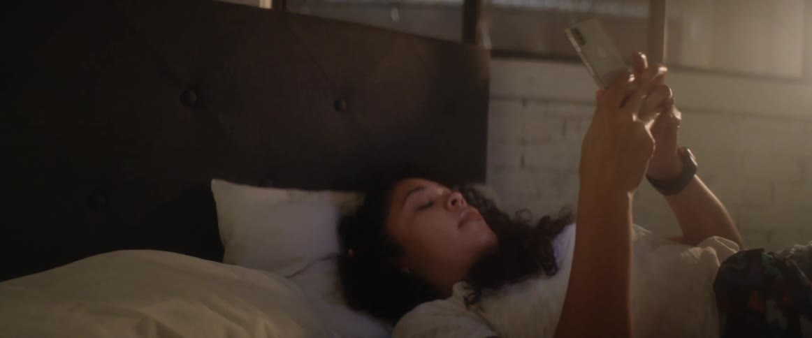 A teenage girl lying in bed scrolls through her cell phone