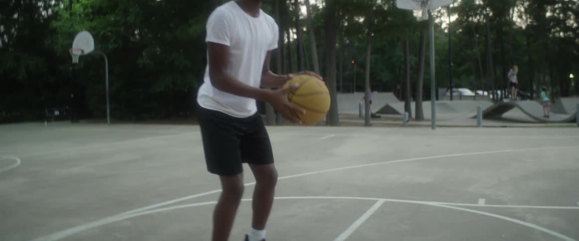 A black teenage male stands at a free throw line on an outdoor basketball court as he catches and shoots a basketball