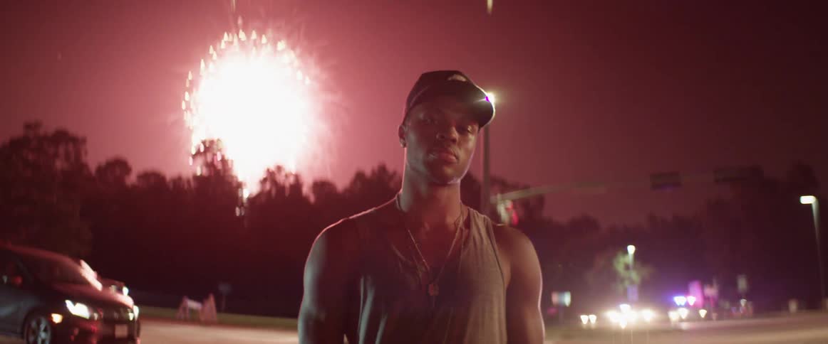 A man with a hat standing with vehicles and fireworks in the sky in the background