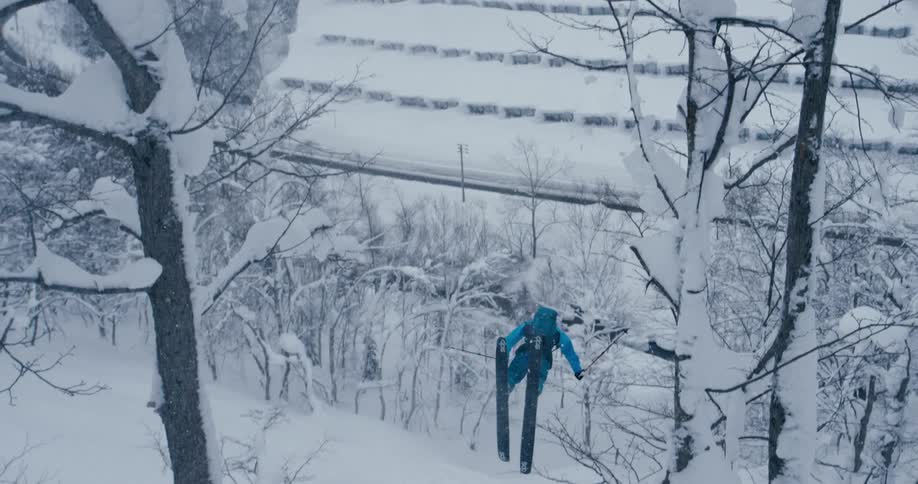 A person with a helmet and a backpack skiing and flipping down a snowy slope with trees