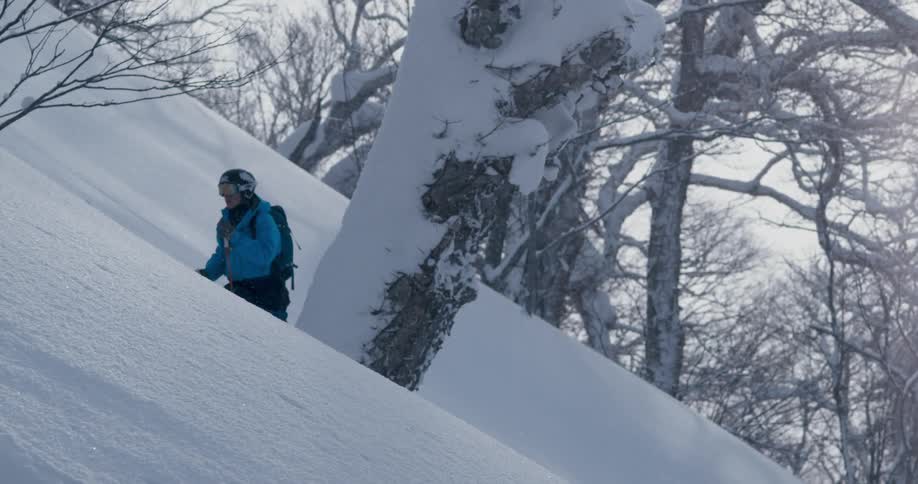 A person with a helmet and goggles and a backpack going up a snowy slope with trees