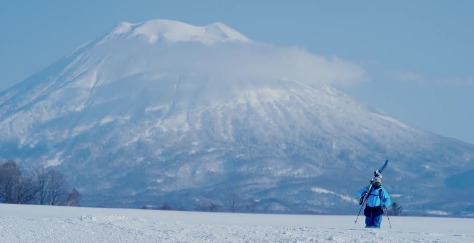 A person with a hat and coat walking with skis on their back with a mountain in the background