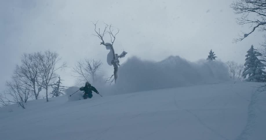 A person with a coat gloves and goggles skiing down a snowy slope with trees