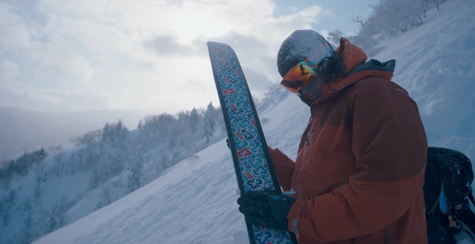 A skier standing on a snowy mountain slope wipes clean the bottom of a ski