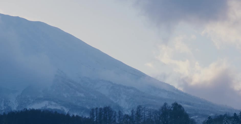 A massive mountain is covered in snow as thick clouds pass nearby to it