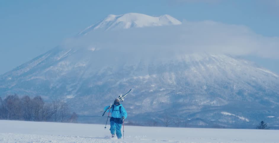 A hiker hikes through a field of snow beneath a snow capped mountain during the day