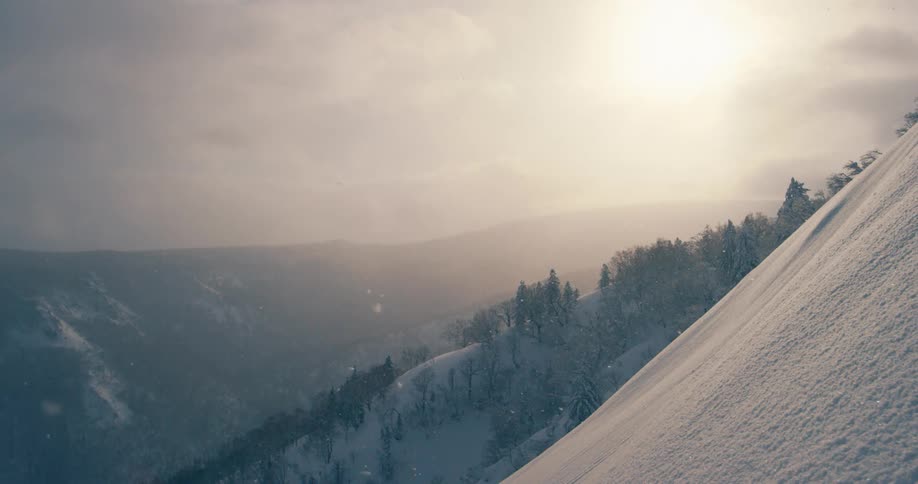 Snow falls onto steep snow covered slope near forest on cloudy day