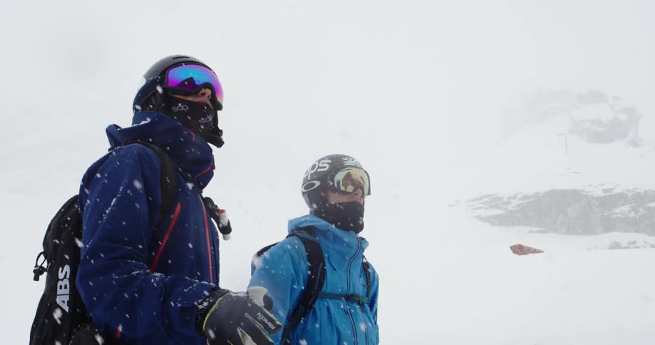 Two people wearing cold weather gear stand in snowstorm with snow covered slope in background
