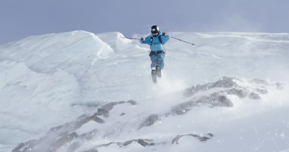 A skier in a blue coat going over a jump in snowy mountains