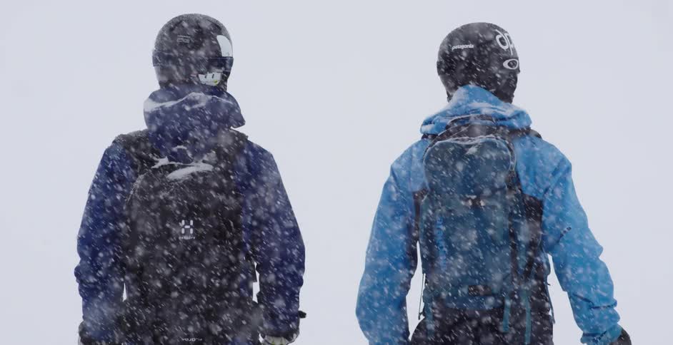 Two people stand wearing cold weather gear and helmets as snow falls