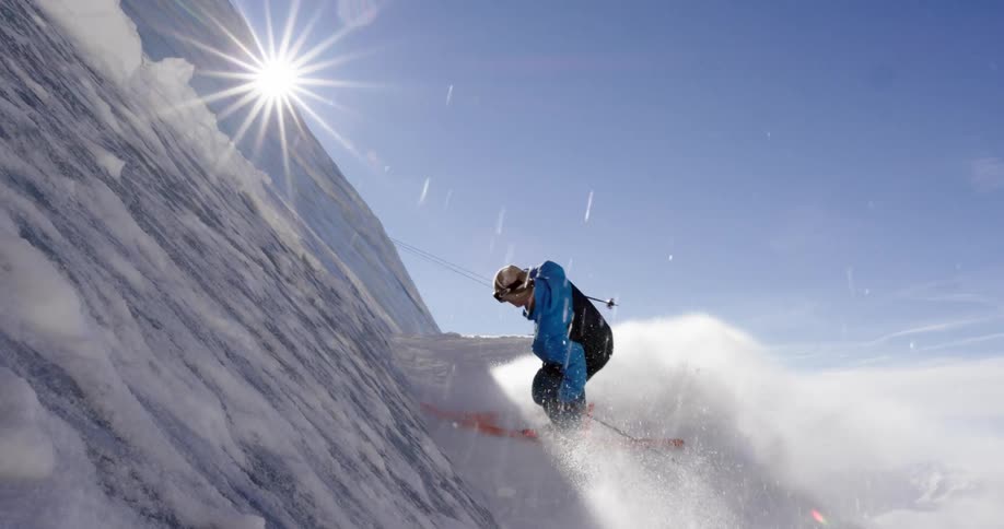 A skier slides to a stop on a ledge on a snow covered mountain on a sunny day
